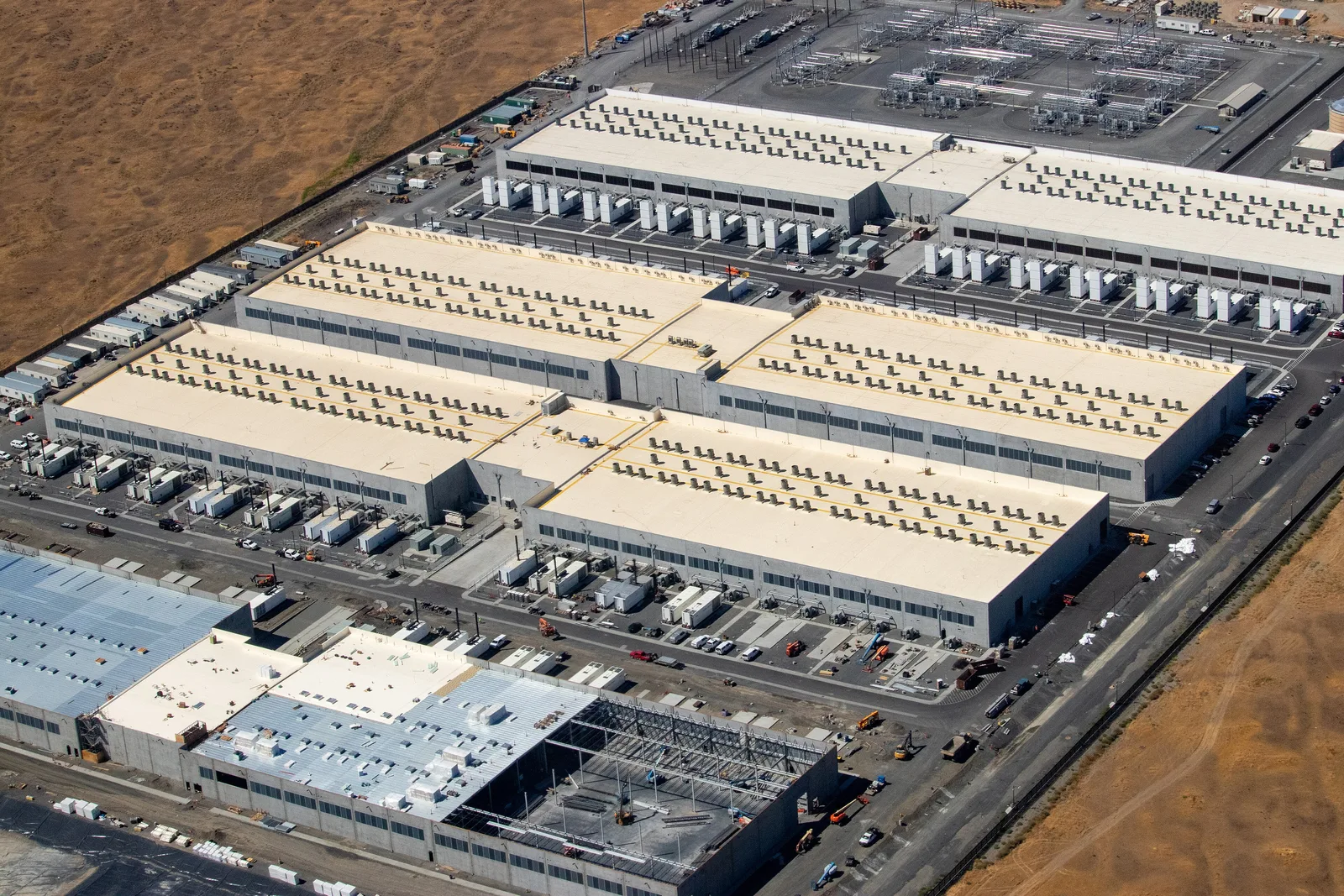 Aerial view of three large data center buildings with a fourth under construction, surrounded by cooling units and a power substation
